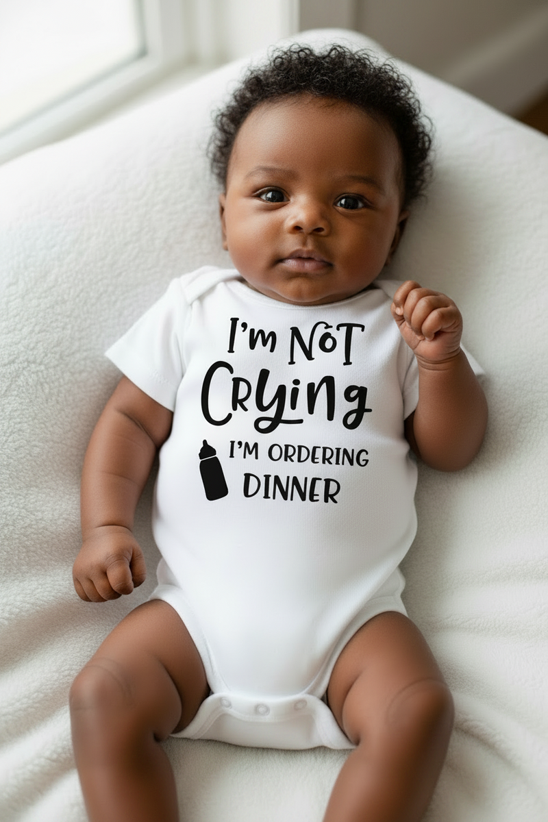 Baby wearing a white onesie with text sitting on a white blanket.